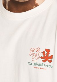 Vit bomull t-shirt med ett grafiskt mönster. Innehåller två stiliserade växter i röd och en grön "Quiksilver" text med "Tripping Since '69."