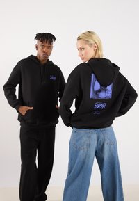 Black hooded sweatshirts feature front pockets and graphic designs on the back. One model wears black pants; the other wears blue jeans.