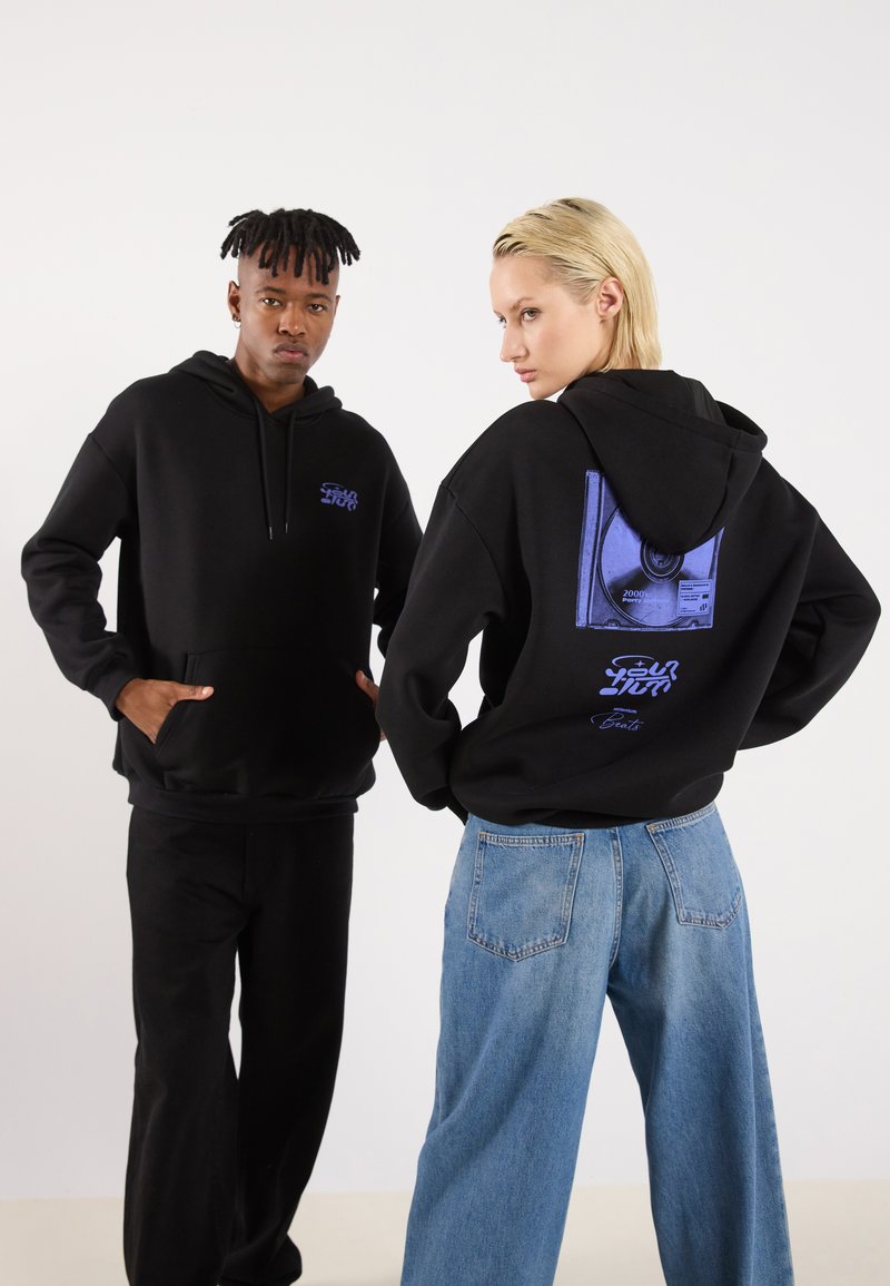 Black hooded sweatshirts feature front pockets and graphic designs on the back. One model wears black pants; the other wears blue jeans.