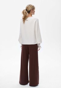 Witte textuurpullover met pofmouwen, gecombineerd met brede bruine broek. Opvallende details zijn de franje aan de mouwen en een klein logo op de achterkant.