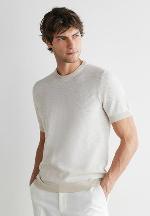 Man met bruin haar die een beige kortemouwen gebreide trui en een witte broek draagt, staat met zijn handen in zijn zakken en kijkt opzij.