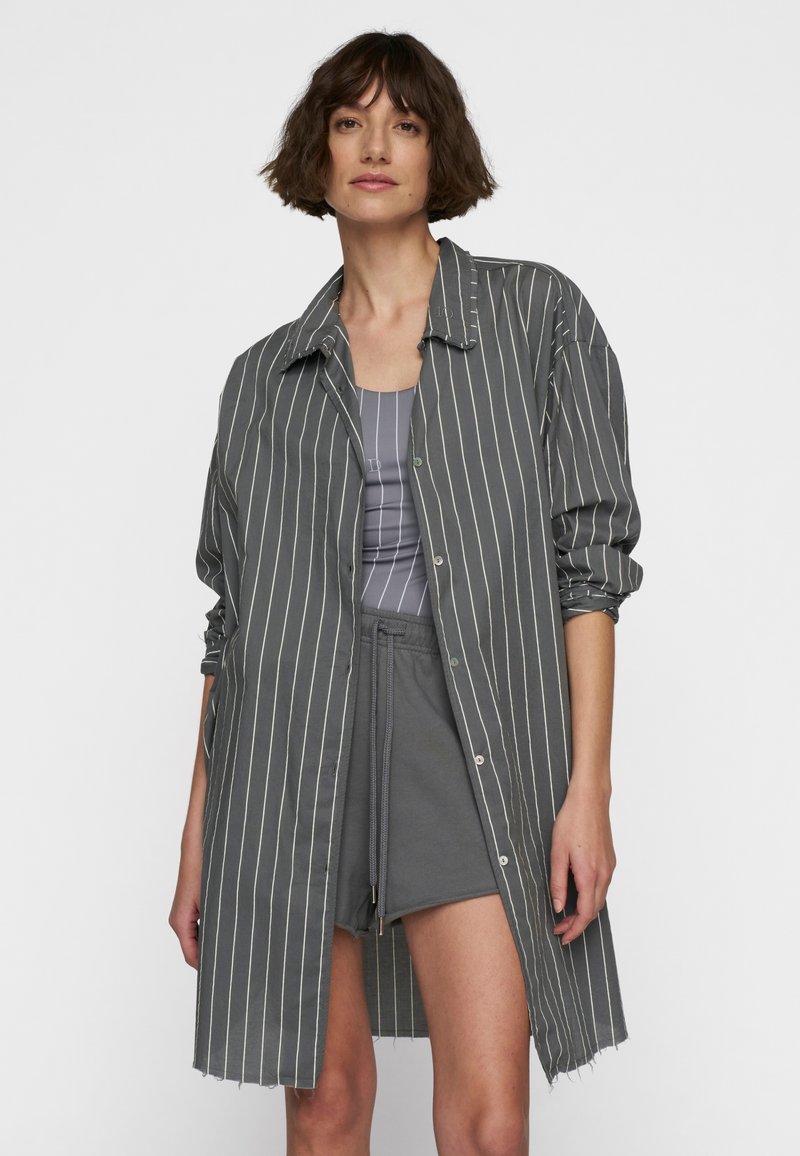 Camicia a righe grigie con bottoni sul davanti, maniche lunghe arrotolate e un orlo asimmetrico. Indossata sopra un top grigio e pantaloni corti.