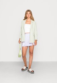 Blazer verde claro, top branco cropped, saia mini lilás e sandálias pretas com detalhes brancos. A bolsa é de um verde texturizado. Fundo suave.
