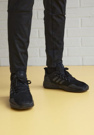 Personne portant des chaussures de sport noires et un pantalon noir, debout sur un sol jaune devant un mur en briques blanches.
