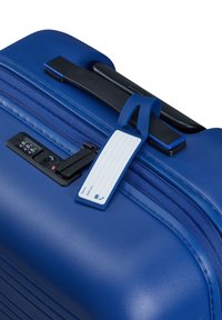 Valise rigide bleue avec une surface texturée, une poignée noire, un cadenas à combinaison et une étiquette de bagage bleue avec un champ d'adresse blanc.