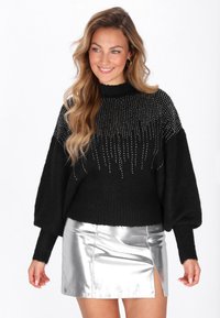 Femeie zâmbind, îmbrăcată cu un pulover tricotat negru cu cusături verticale argintii și o fustă mini argintie lucioasă cu o deschidere în față, stând în fața unui fundal alb.