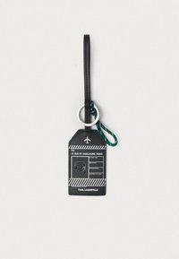 Fekete poggyász címke fehér utazási információkkal, napszemüveget viselő rajzolt arc, zöld hurok és "Karl Lagerfeld" márkajelzés világos háttéren.