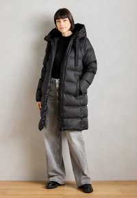 Cappotto imbottito nero, con colletto alto, chiusura con zip e cappuccio. Presenta sezioni trapuntate, tasche laterali ed è abbinato a jeans grigi a gamba larga.
