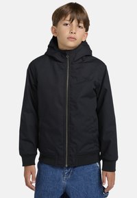 Sort hættejakke med frontlynlås, blød tekstur, elastiske manchetter og sidelommer, båret over denim. Subtil branding på brystet.
