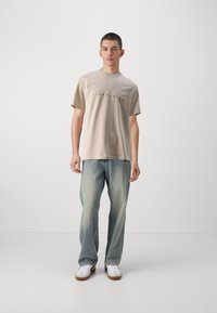 Camiseta de algodón beige con cuello redondo y texto impreso en negro. Combinada con jeans azules claros y zapatillas blancas con suelas marrones.