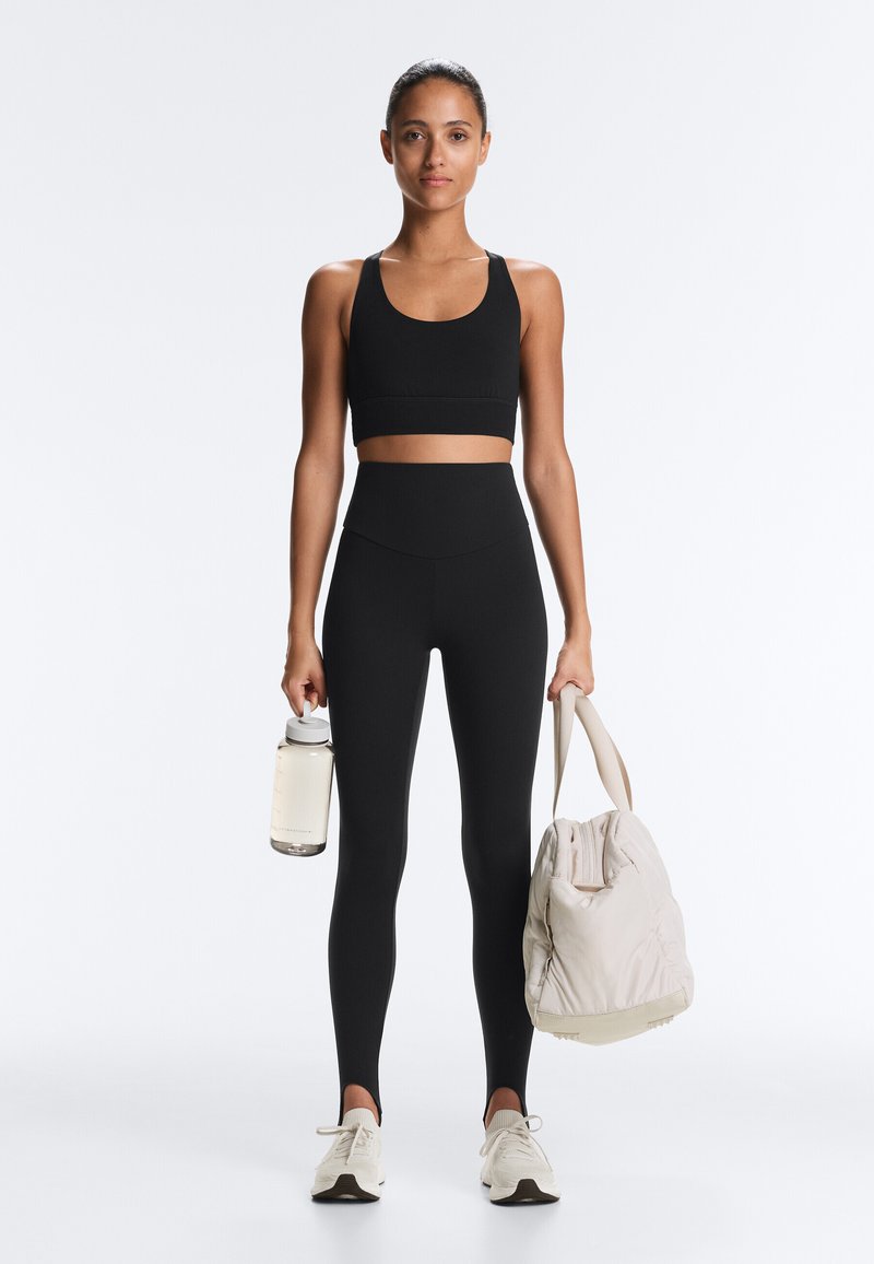 Vrouw in een zwarte sport-bh en legging, met in de ene hand een waterfles en in de andere hand een beige sportschooltas, staand tegen een effen achtergrond.