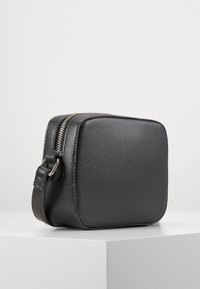 Borsa a tracolla in pelle nera texture con forma rettangolare, chiusura superiore con zip e cintura regolabile sottile. Design minimalista senza loghi visibili.