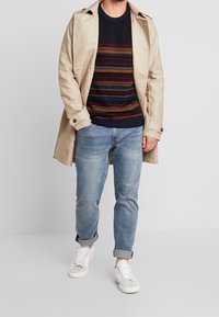 Manteau trench beige sur un pull à motifs bleu marine et multicolores, associé à un jean léger bleu clair retroussé et des baskets blanches.