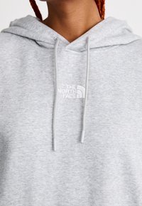 Personne portant un sweat à capuche gris clair de North Face avec des cordons de serrage visibles et un logo sur la poitrine, avec des cheveux tressés partiellement visibles.