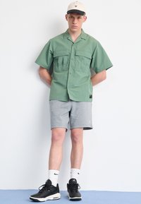 Grön kortärmad skjorta med två framfickor, kombinerad med grå shorts. Svarta sportiga sneakers och en beige keps med svart brätte.