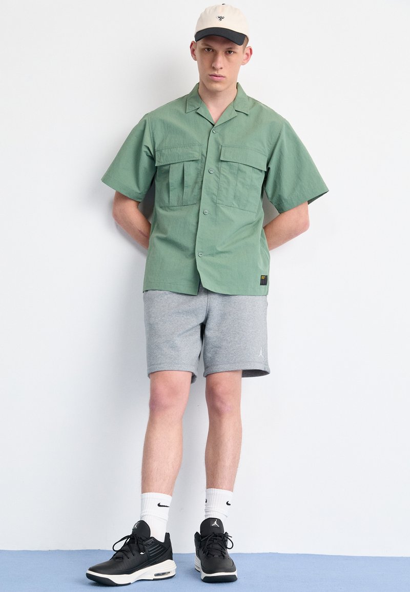 Camisa verde de manga corta con dos bolsillos frontales, combinada con pantalones cortos grises. Zapatillas deportivas negras y una gorra beige con visera negra.