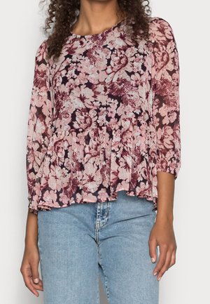 Woman wearing a pink and burgundy floral long-sleeve blouse with a ruffled hem, paired with light blue denim jeans.