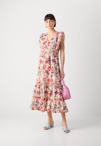 Rochie maxi floral în culori pastelate, cu mâneci încrețite, tiv în straturi și talie strânsă. Asortată cu o geantă roz și mules albastre.