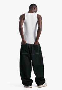 Moški v beli majici brez rokavov in prevelikih črnih hlačah z vidnimi zadnjimi žepi, stoji z rokami v žepih pred belim ozadjem.