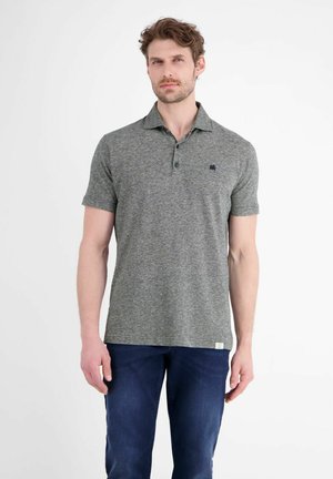 Mann trägt ein graues kurzärmeliges Poloshirt und dunkelblaue Jeans, steht vor einem schlichten weißen Hintergrund.