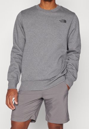 Mann trägt ein graues Sweatshirt mit Rundhalsausschnitt und The North Face-Logo sowie graue Freizeithosen, steht mit einer Hand in der Tasche.