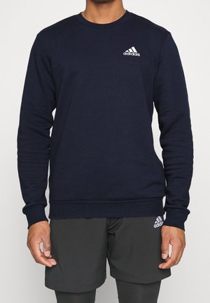 Osoba ubrana w granatową bluzę Adidas i czarne spodenki Adidas, oba z białym logo Adidas po lewej stronie.