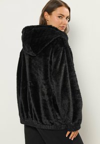 Czarna kurtka z sztucznego futra z kapturem, o miękkim, teksturowanym wykończeniu, luźnym kroju i elastycznym dołem. Posiada zaokrąglony kaptur i długie rękawy.