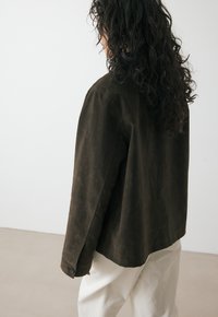 Veste en daim marron avec une coupe décontractée, des poignets boutonnés et une texture mate. Présente un design minimaliste et des lignes épurées.