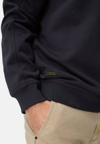 Sweat-shirt bleu marine foncé avec une finition texturée, arborant une petite étiquette rectangulaire indiquant "FABRIQUÉ POUR LES AVENTURES QUOTIDIENNES", associé à un pantalon kaki.