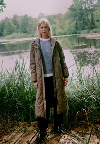 Cămașă lungă, căptușită de culoare khaki, cu un model matlasat, purtată peste un pulover gri cu dungi și pantaloni negri, având un fundal natural.