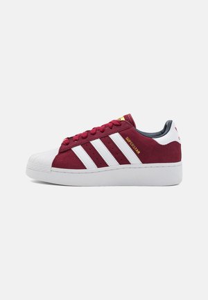 Bordeauxrote und weiße Adidas Superstar Sneaker mit weißer Shell-Toe, drei weißen Streifen, goldenem "Superstar"-Schriftzug und weißer dicker Sohle.