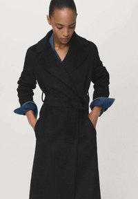 Manteau trench en laine noire avec un design enveloppant, un large col et des manches longues. Présente des accents en denim au niveau des poignets et une ceinture à nouer.