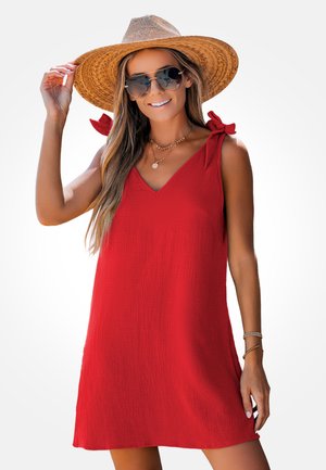 Femme portant une robe rouge sans manches avec des bretelles nouées aux épaules, un chapeau de paille à large bord marron, des lunettes de soleil et des colliers dorés superposés, souriant.