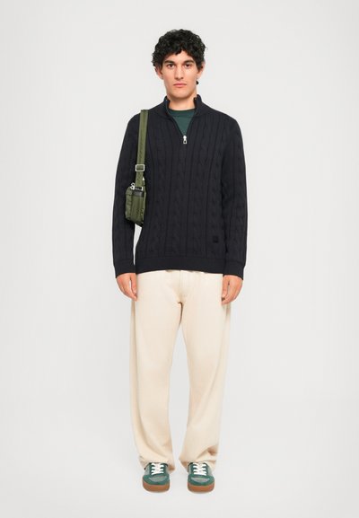 Νεαρός άνδρας φοράει ένα μαύρο ζακέτα με πλεχτό σχέδιο, μπεζ παντελόνια, πράσινα αθλητικά παπούτσια και κρατάει μια ελαιόχρωμη τσάντα χεριού σε λευκό φόντο.