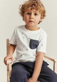 Weißes, geripptes T-Shirt mit kurzen Ärmeln, ausgestattet mit einer dunkelblauen Tasche und kleinen bedruckten Booten. Kombiniert mit strukturierten dunkelblauen Hosen.