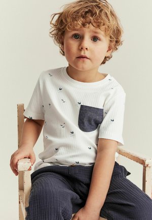 Weißes, geripptes T-Shirt mit kurzen Ärmeln, ausgestattet mit einer dunkelblauen Tasche und kleinen bedruckten Booten. Kombiniert mit strukturierten dunkelblauen Hosen.