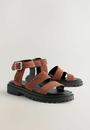 Brązowe zamszowe sandały z wieloma szerokimi paskami, srebrnymi ćwiekami i czarną grubą podeszwą. Zapięcie akcentujące na pasku kostki.