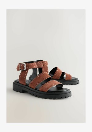 Brązowe zamszowe sandały z wieloma szerokimi paskami, srebrnymi ćwiekami i czarną grubą podeszwą. Zapięcie akcentujące na pasku kostki.