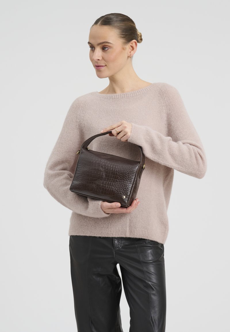 Femme en pull beige et pantalon en cuir noir tenant un sac à main marron foncé à texture crocodile avec poignée supérieure.