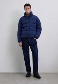 Hombre con una chaqueta acolchada azul marino sobre una camiseta gris, pantalones vaqueros azul oscuro y zapatos negros, de pie contra una pared clara y sencilla.