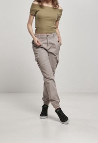 Top off-shoulder color oliva chiaro abbinato a pantaloni cargo beige con polsini elasticizzati e tasche laterali, completato da sneakers nere.
