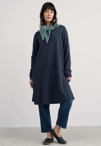 Nainen, jolla on päällä pitkä tummaa harmaata mekkoa sinisten farkkujen päällä, mustat liukuvarvas kengät ja vaaleansininen neulottu huivi kaulassa.