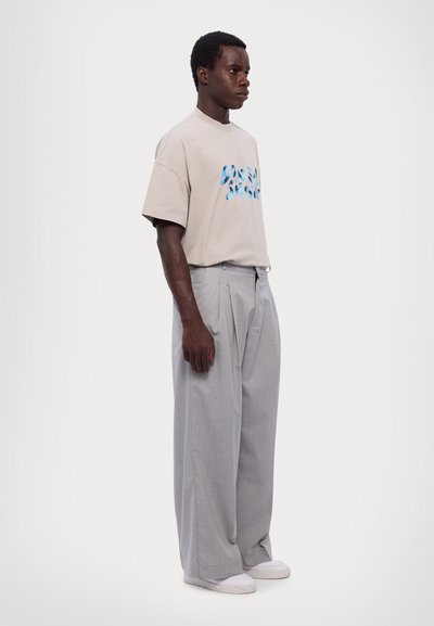 Beige cotton t-shirt with blue graphic design, paired with light gray, pleated dress pants, and white slip-on shoes.