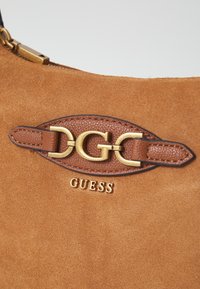 Ruskea mokkanahkainen käsilaukku, jossa on kultainen metallikoriste, jossa on toisiinsa lomittuvat kirjaimet "DGC" ja alapuolella kohokuvioitu brändinimi "GUESS".