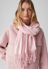 Écharpe en cachemire rose à bords frangés, portée lâchement autour du cou, associée à un sweat-shirt rose assorti avec une poche.