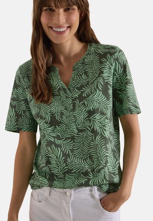 Femme portant une chemise verte avec un motif de feuille de palmier vert clair et un pantalon blanc, souriante avec une main dans la poche.