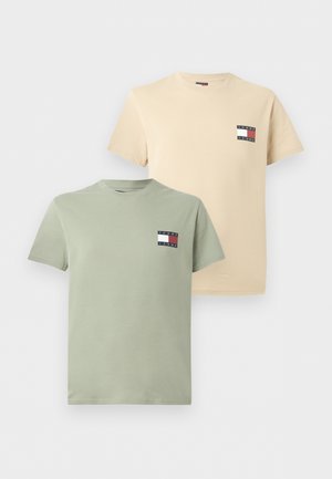 Man met handen in de zakken, draagt een beige T-shirt met korte mouwen en logo, losse blauwe jeans en bruine sneakers, kijkt iets naar rechts.