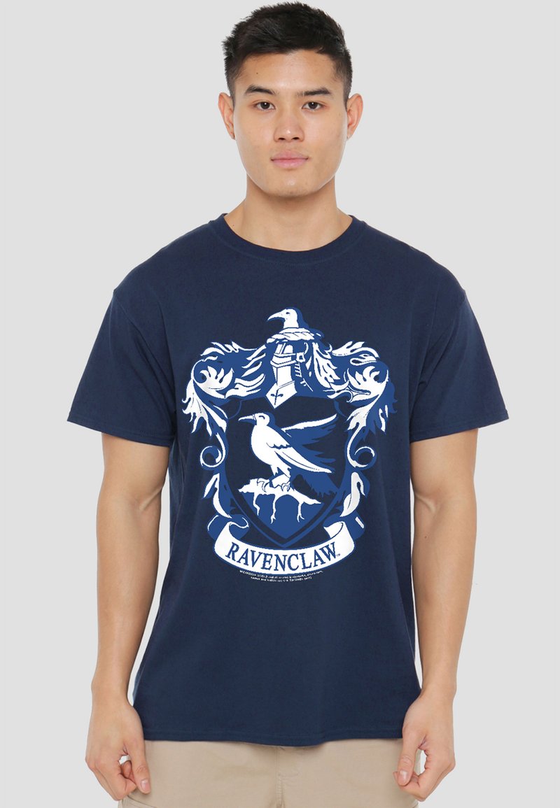 Young man wearing navy blue T-shirt with white Ravenclaw house crest and beige shorts, standing against plain light background.