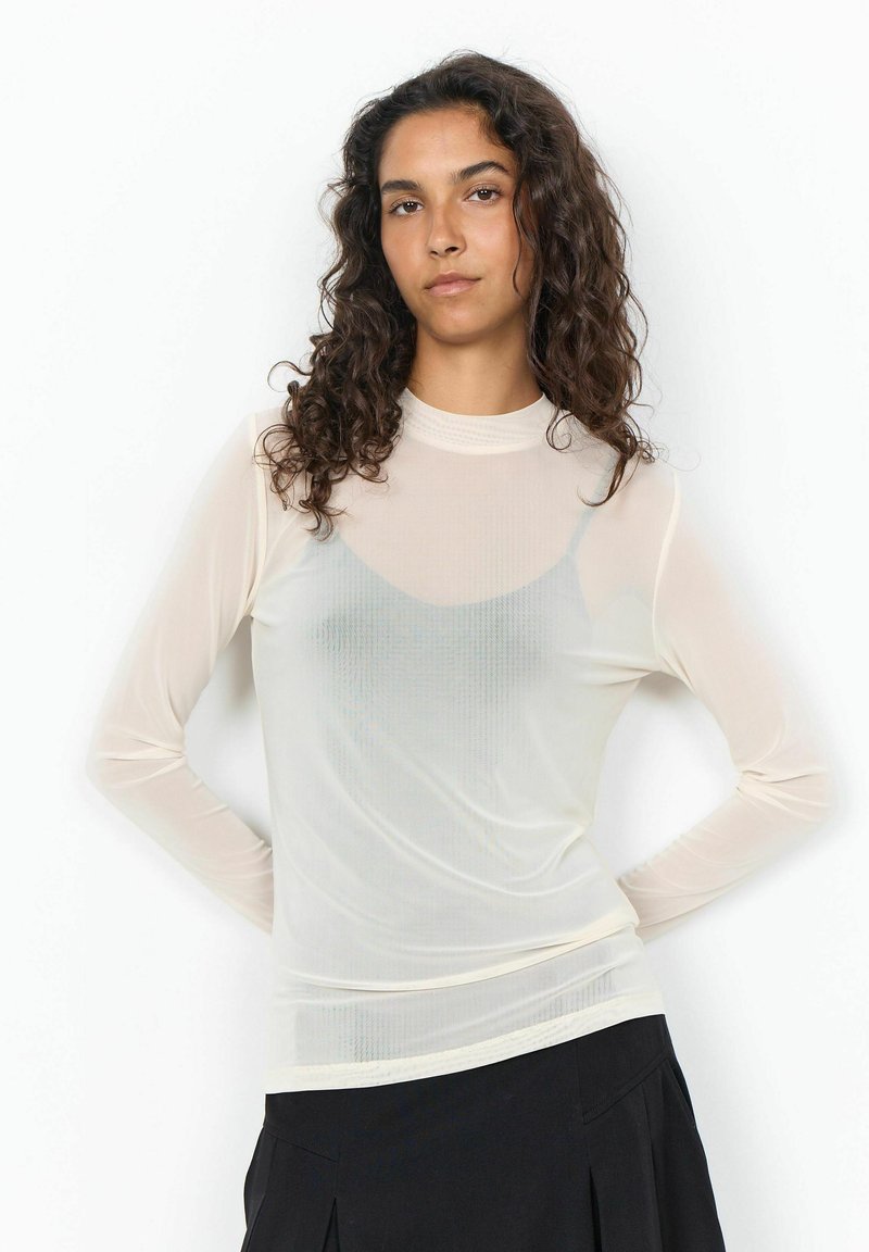 Femme aux cheveux bouclés foncés portant un haut blanc transparent à manches longues par-dessus un débardeur gris clair et une jupe noire, debout, les mains sur les hanches.