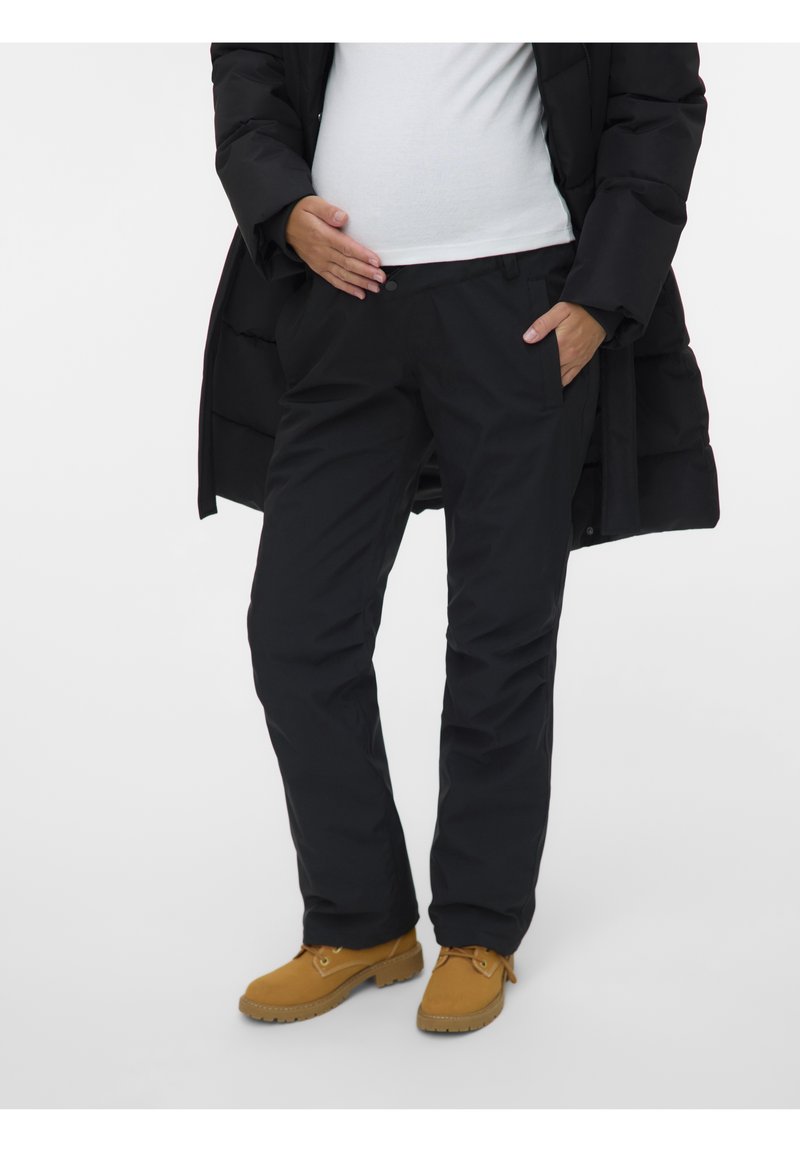 Personne portant un pantalon noir, une chemise blanche, un manteau noir et des bottes beige, avec une main dans la poche et l'autre reposant sur le ventre.